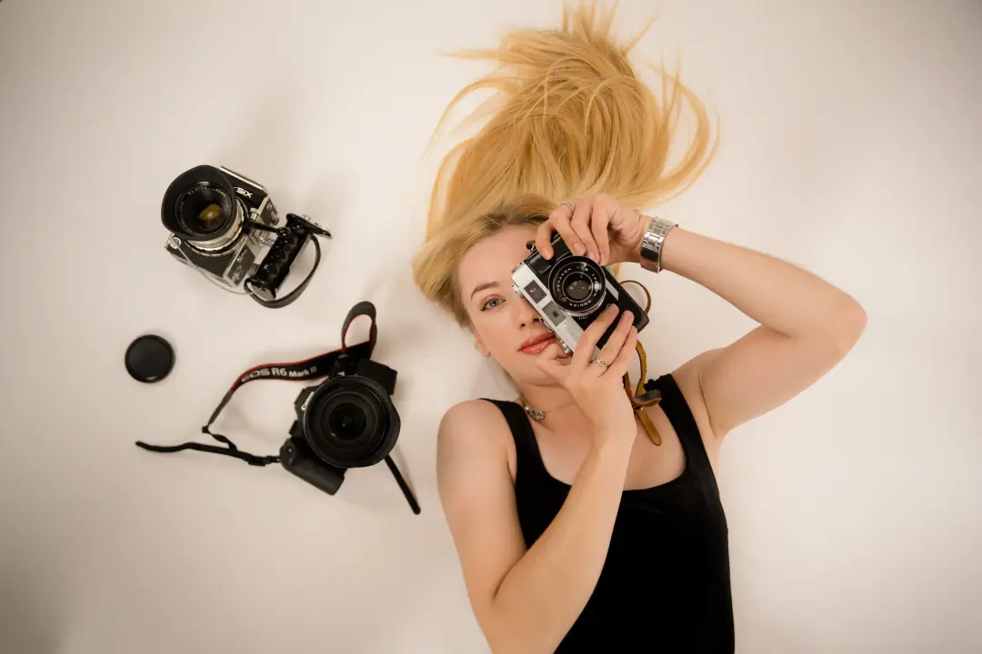 A blonde photographer lies on a light backdrop looking through a vintage film camera, with two more cameras and a lens arranged beside her.